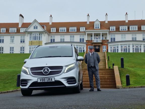 Man standing next to silver Mercedes in front of a large white hotel building with red roof.