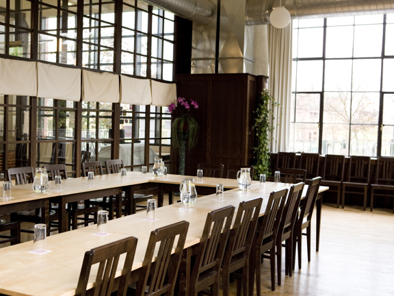 A room set up at the West brewery for a private meeting. Several pale wood tables are arranged into a U-configuration. The outside edge is lined with dark wooden, upright chairs, glasses and water jugs sit on the table tops. The floor is polished wood. The left hand wall is glass, made up of paned panels. The right-hand exterior wall is a large, landscape window with cream curtains pulled back. Spare chairs are lined up under the window. Potted plants and minimal spherical ceiling lights decorate the room.