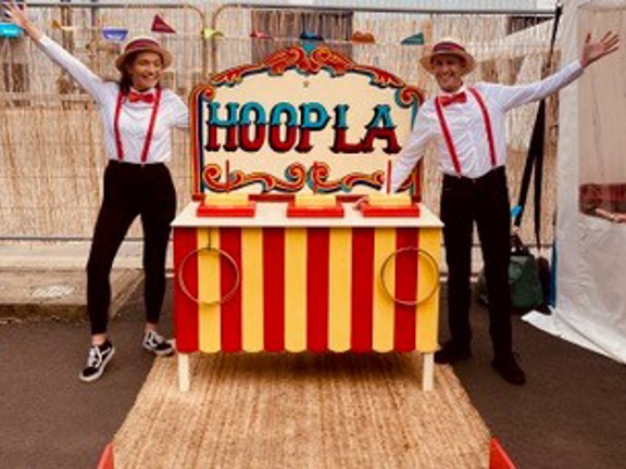 People standing next to an old fashion circus stall that reads Hoopla