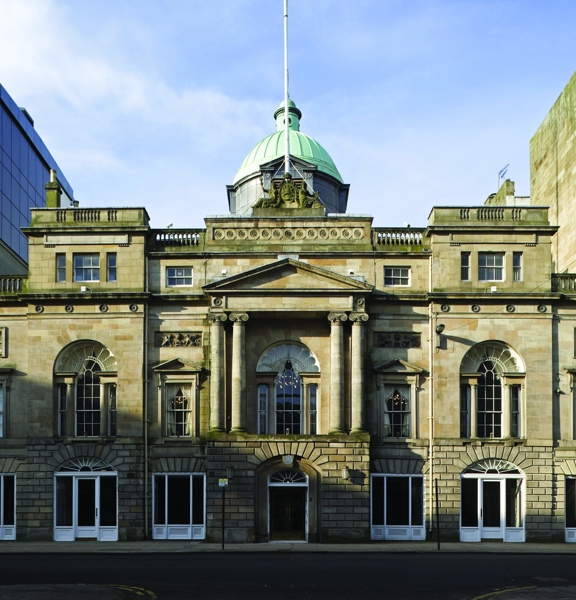 An exterior of the Glasgow Trades Hall shows a 3 storey, symmetrical, Victorian sandstone building. It has carved details, including ionic columns either side of the central, 1st floor arched window and a carved coat of arms finial on top. A large verdigris dome and a flagpole also adorn the top of the building. The image is taken from over the road, a level pavement can be seen outside of the building's entrance. Buildings flank the Trades Hall on either side.