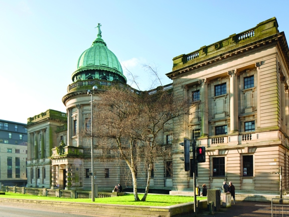 An exterior view from the street outside the Mitchell Library Complex. Shows a pedestrian crossing outside a large 3-storey sandstone institution. A verdigris dome topped with a female figure, adorns the central curving portion of the building, above a large, pillared doorway. In the foreground, a grassy bank with a silver birch in its centre sits slightly raised from the tarmac pavement around it.  Lampposts and a modern building can be seen in the background.
