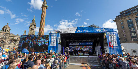 Crowds gathered in front of blue stage with screen showing medals being presented in George Square, Glasgow.
