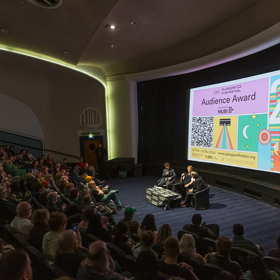 Three people sit in front of a busy audience in the GFT cinema. The screen says  'Audience Award'.