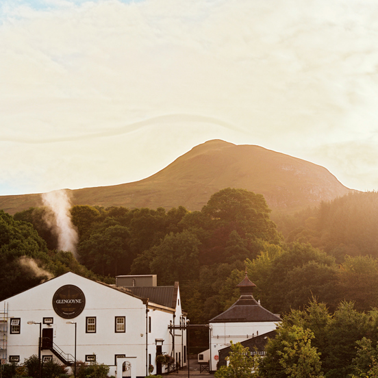 An exterior view of the Glengoyne Distillery shows a 3 storey building with white rendering and a gabled roof, dwarfed by the trees and hills behind it. There is a neighbouring smaller building obscured by trees. The large building has a large black circle on it with white lettering inside it reading "Glengoyne." The sky is bright giving a golden glow to the large trees, the striking outline of a hill and the sky. It also highlights the steam or smoke rising from the main building. 