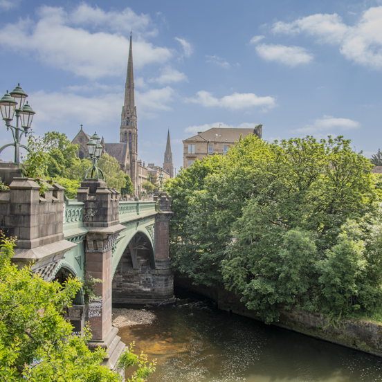 Sunny view of the green metal Kelvin Bridge surrounded by greenery and a tall church spire in the background