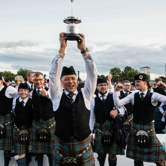 A piper grins and holds a trophy above his head at the World Pipe Band Championships in Glasgow Green.