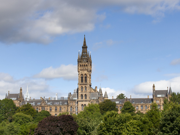 Sunny view of the Gothic revival-style spire of the University of Glasgow's main building, with green trees in the foreground