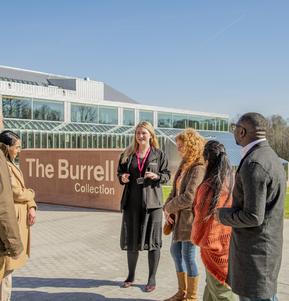 Group of people standing outside a modern red brick and glass building, a museum known at The Burrell Collection. Sun is shining, blue sky and a brick sign behind the group reads The Burrell Collection 