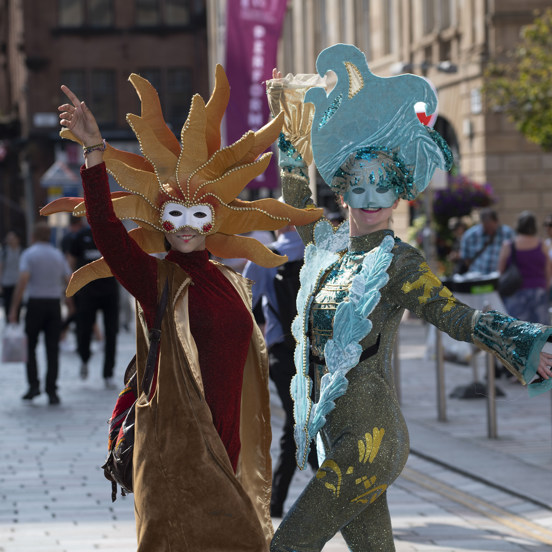 Two performers in carnival costumes pose with their arms up in the street in Glasgow.