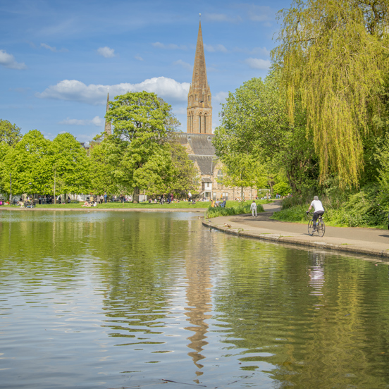 Sunny view of a small, tree-lined pond in Queen's Park, with cyclists and people strolling along a path, with a church spire in the background