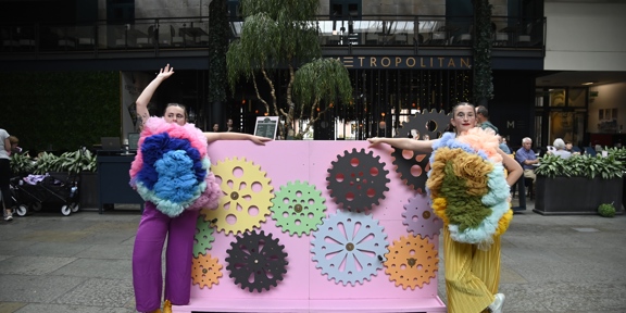 Two performers in pom-pom costumes stand in front of a display of colourful cogs in Merchant Square, Glasgow.