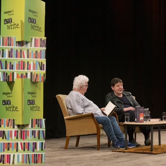 Two authors sit in armchairs on stage, with a green Aye Write banner on each side.