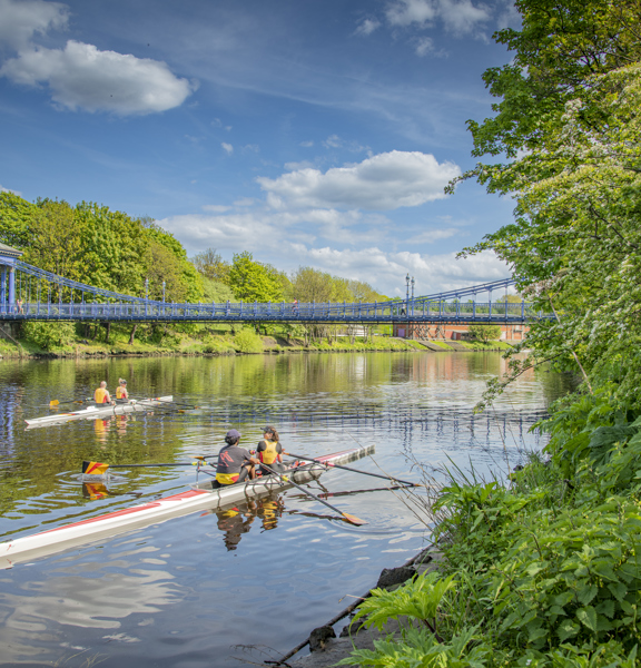 Sunny view of 4 rowers on 2 double sculls on the tree-lined River Clyde with the blue St Andrew's suspension footbridge in the background