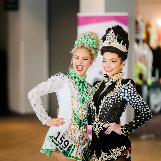 Two Irish dancers in a black dress and a white and green dress smile while having their photo taken.
