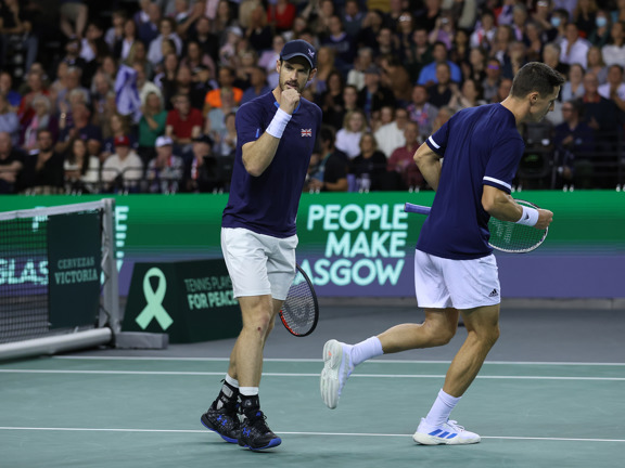 Two male tennis players wearing navy uniforms stand on court, in front of a People Make Glasgow sign.
