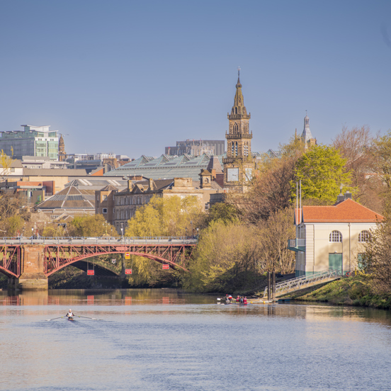 Sunny view of rowers on single sculls on the tree-lined River Clyde with a bridge and the city in the background