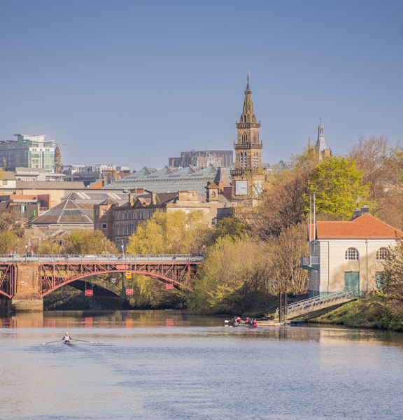 Sunny view of rowers on single sculls on the tree-lined River Clyde with a bridge and the city in the background