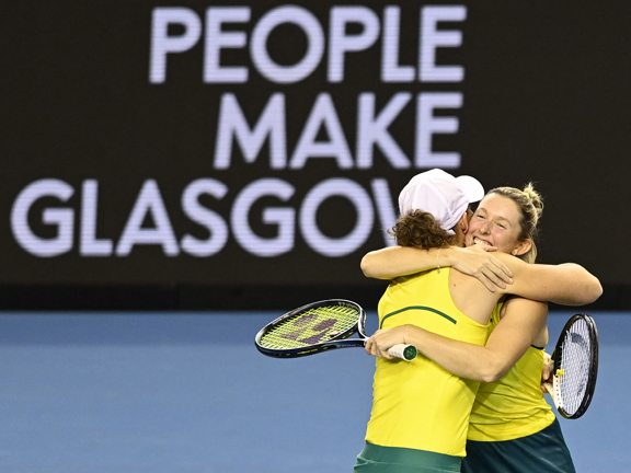 Two women tennis players, wearing yellow, hug on court in front of a People Make Glasgow sign.