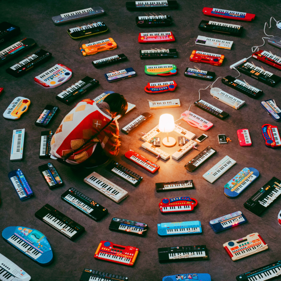 Person crouches in front of a lamp, surrounded by different kinds of portable music keyboards that are arranged in a circle.