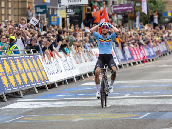 Woman cyclist crosses race finish line with hands on head, in front of cheering crowds in Glasgow.