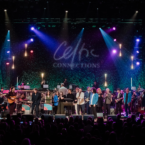 A large group of musicians perform on stage in front of Celtic Connections logo.