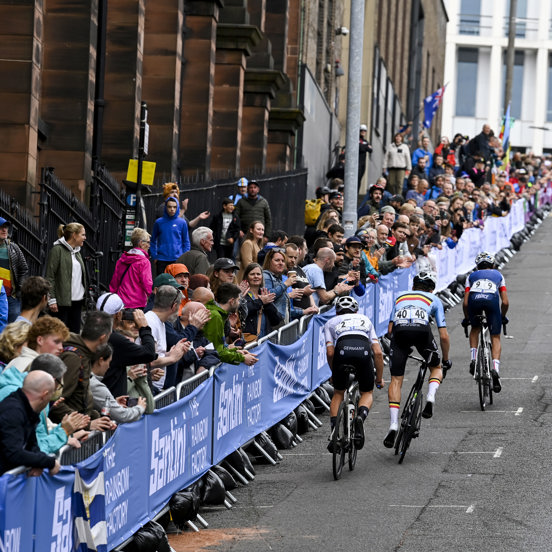 Four cyclists cycle up a steep hill in Glasgow city centre, with a small crowd cheering beside them.
