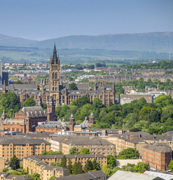 Sunny cityscape with the Gothic revival-style spire of the University of Glasgow's main building, surrounded by greenery and buildings of the West End, including the Baroque-style Kelvingrove Art Gallery and Museum