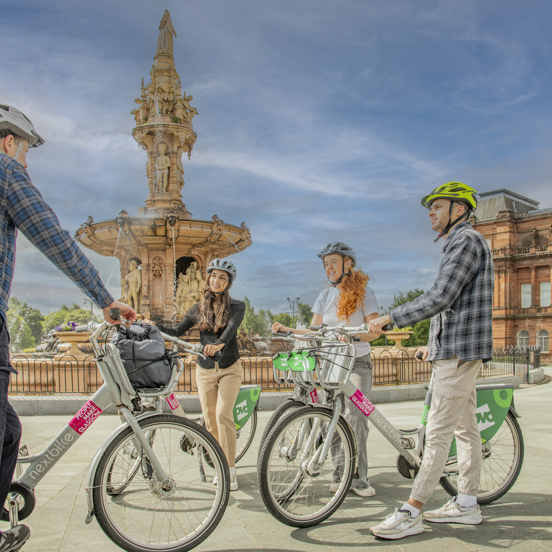 4 cyclists chatting in front of the large terracotta, five-tier, French Renaissance-style Doulton Fountain in Glasgow Green, with the later French Renaissance-stye People's Palace in the background