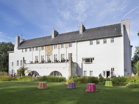 Art nouveau-style white building of the House for an Art Lover, set in a green Bellahouston Park with people socialising on the terrace and with colourful large plastic upside down cake forms on the grass in front