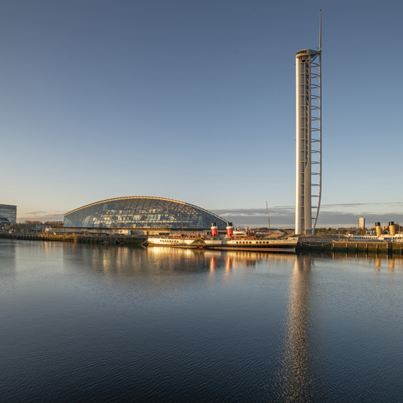 View across the River Clyde of the square, glass building of BBC Scotland, of the crescent-shaped glass front of the Glasgow Science Centre and of the tall metal spire of the Glasgow Tower, with the Waverley paddle steamer docked in front