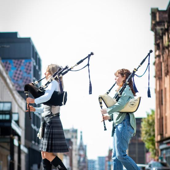 Two bagpipers walk along a sunny Glasgow street, one wear traditional Scottish clothing and one wears casual contemporary clothes.