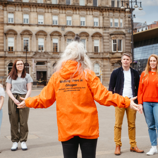 Four individuals, 3 women and one man, stand facing a tour guide with long white hair in a bright orange jacket. The tour guide has her back to us and her arms spread, expressively. Her jacket has black writing, obscured partially by her hair, it is possible to read "Walking Tours Glasgow". The group are stood on an expanse of paved ground. Tall sandstone buildings, a modern glass building, benches and a bronze statue on a plinth are visible behind them.