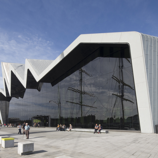 Sunny view of the modern, glass-fronted Riverside Museum, where the roofline reminiscent of a heartbeat on a monitor, with people enjoying the sunshine. The reflection of tall ship masts can be seen in the glass front of the museum