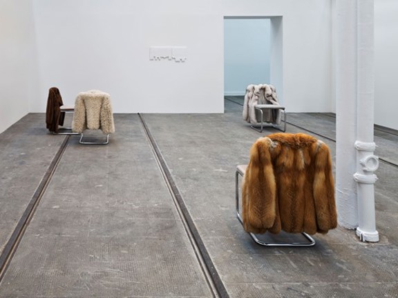 Four metals with fur coats hanging on the back are arranged in a gallery space with white walls.